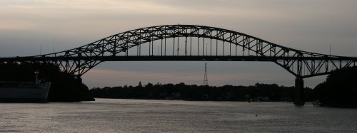 Silhouette of a large bridge spanning a river at dusk, with a calm water surface and distant trees.