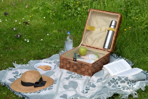 A picnic setup with a woven basket, a water bottle, an apple, cookies, a book, and a straw hat on a grassy field.