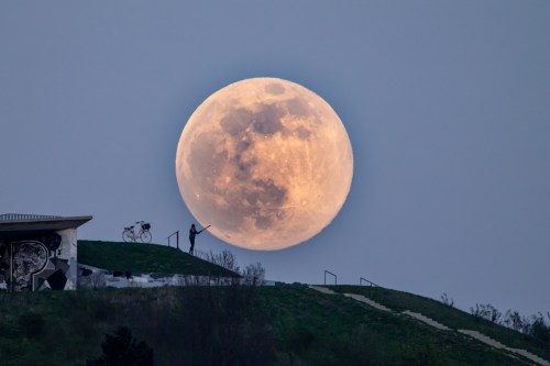 A person stands on a hill, reaching towards a large, glowing full moon in the evening sky. A bicycle is nearby.