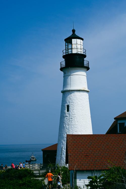A tall white lighthouse stands by the ocean, with people nearby and a clear blue sky above.