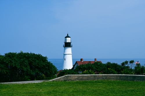 A white lighthouse stands near the ocean, surrounded by greenery and a clear blue sky.