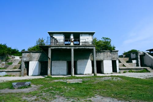 Abandoned concrete building with a flat roof, surrounded by greenery and steps leading up to it under a clear blue sky.