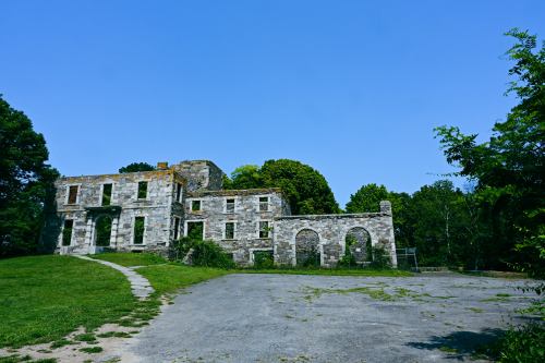 Ruins of a stone building surrounded by greenery under a clear blue sky. Pathway leads to the structure.