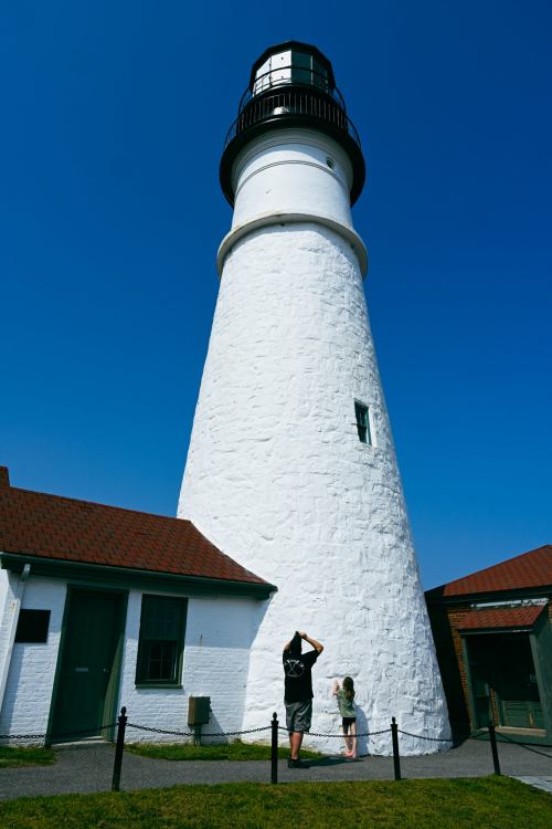 A person and a child stand near a tall white lighthouse against a clear blue sky.