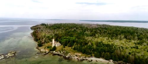 Aerial view of a lush, green island with a lighthouse and rocky shoreline, surrounded by calm waters.