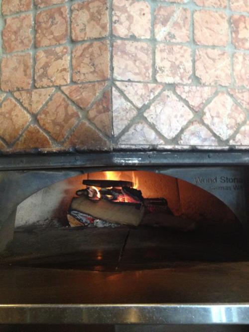 A wood-fired oven with flames visible, set against a tiled wall. The oven has a metallic base and a warm glow.