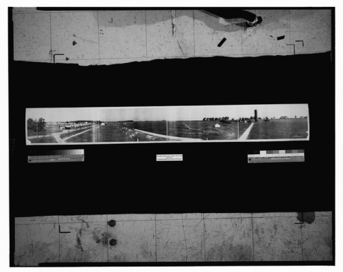 A panoramic black-and-white landscape photo showing fields, buildings, and a distant horizon.