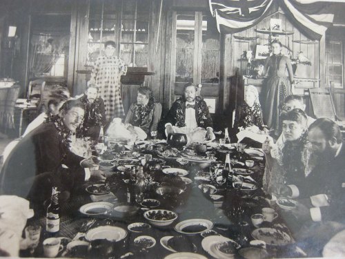 A historical black-and-white photo of a large group dining together, surrounded by food and festive decorations.