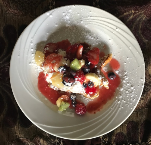 A plate of French toast topped with mixed fresh fruits and a drizzle of berry sauce, dusted with powdered sugar.