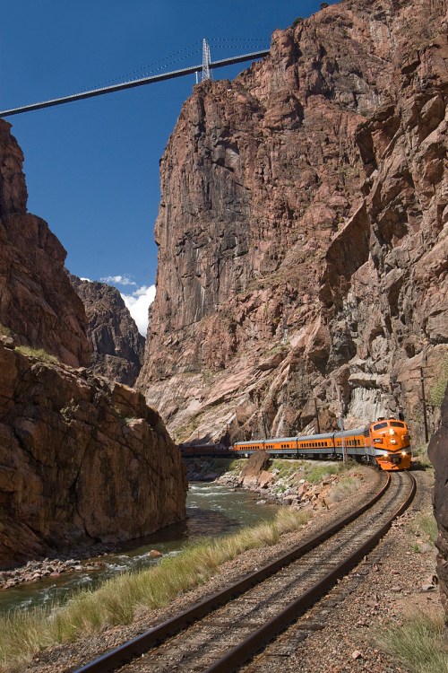 A vibrant orange train curves through a rocky canyon, with a bridge high above and a river flowing nearby.