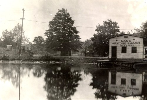 Black and white photo of a lake with a building labeled "Lake Hopatcong Gardens" and trees reflected in the water.