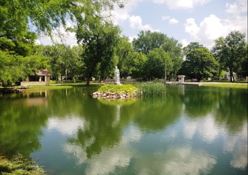 A serene pond surrounded by trees, featuring a statue on a small island and reflections in the water.