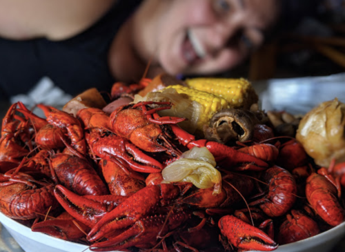 A close-up of a large bowl of crawfish, corn, and other seafood, with a person smiling in the background.