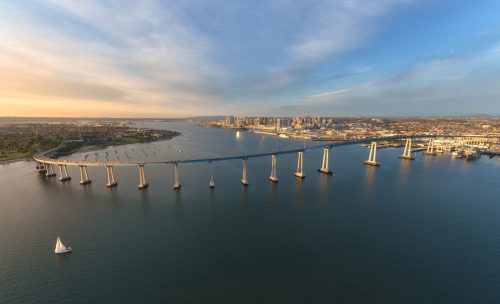 Aerial view of a bridge spanning a calm waterway, with a city skyline in the background and a sailboat in the foreground.
