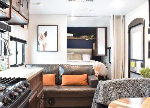 Cozy interior of a camper with a sofa, kitchen area, and a bed in the background, featuring warm decor and natural light.