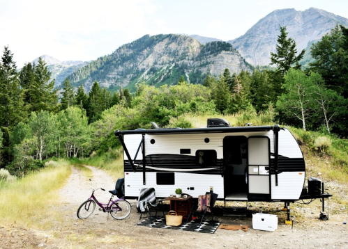 A camper parked on a dirt path surrounded by trees and mountains, with a bike and outdoor setup nearby.