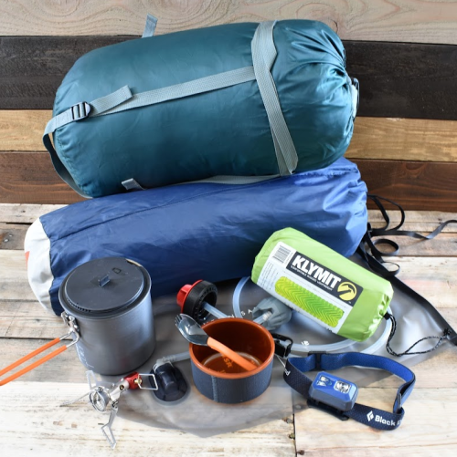 A collection of camping gear including sleeping bags, cookware, utensils, and a headlamp on a wooden surface.