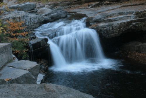 A serene waterfall cascades over rocky ledges into a calm pool, surrounded by autumn foliage.
