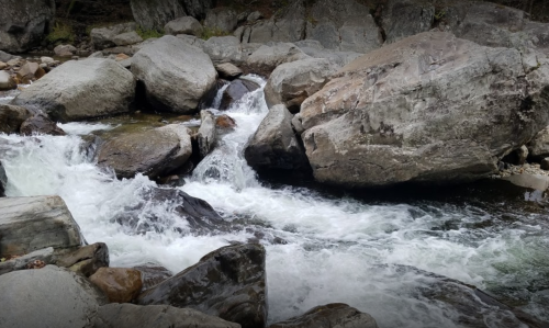 A flowing river cascades over large rocks, surrounded by a natural landscape.