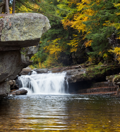 A serene waterfall cascades into a calm pool, surrounded by vibrant autumn foliage and rocky cliffs.