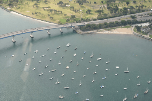 Aerial view of a bridge over water, with numerous boats anchored below and a beach visible nearby.
