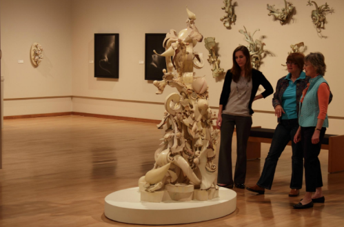 Three women admire a tall, intricate sculpture in an art gallery, surrounded by wall-mounted artworks.