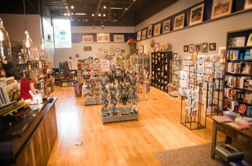 A cozy gift shop filled with various items, including books, art, and decorative pieces, with wooden floors and bright lighting.