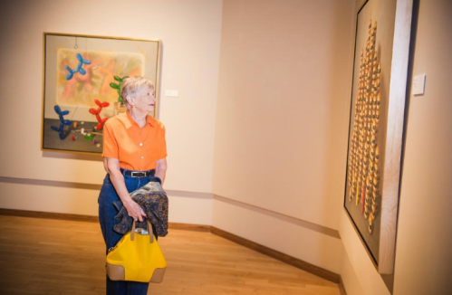 An older woman in an orange shirt admires artwork in a gallery, holding a yellow bag and a scarf.
