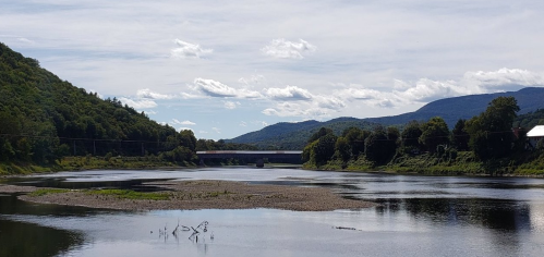 A serene river scene with lush green hills, a bridge in the distance, and a clear blue sky with scattered clouds.