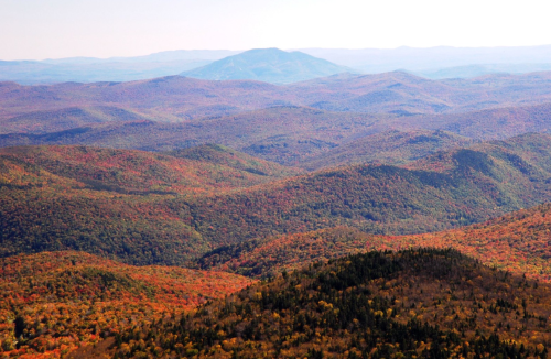 A panoramic view of rolling mountains covered in vibrant autumn foliage, showcasing shades of orange, red, and green.