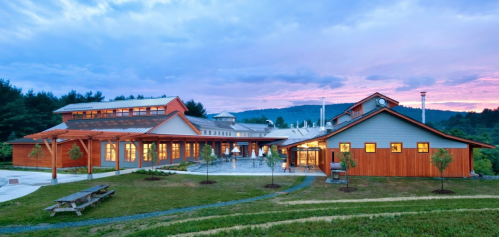 A modern wooden building with large windows, surrounded by greenery and mountains, under a colorful sunset sky.