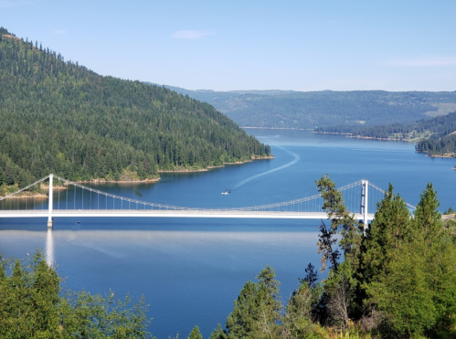 A scenic view of a suspension bridge over a calm lake, surrounded by lush green hills and clear blue skies.