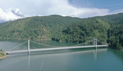 Aerial view of a suspension bridge spanning a calm lake, surrounded by lush green hills and a blue sky.