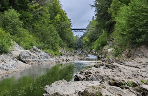 A serene river winding through rocky terrain, surrounded by lush greenery and a bridge in the distance under a cloudy sky.