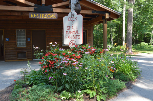 Sign directing to deer and small mammal exhibits, surrounded by colorful flowers near a restroom building in a wooded area.