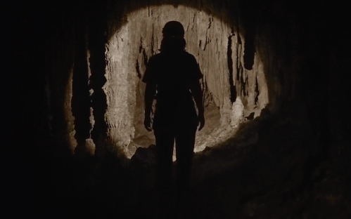 A silhouette of a person standing in a dark cave, illuminated by a soft light from behind.