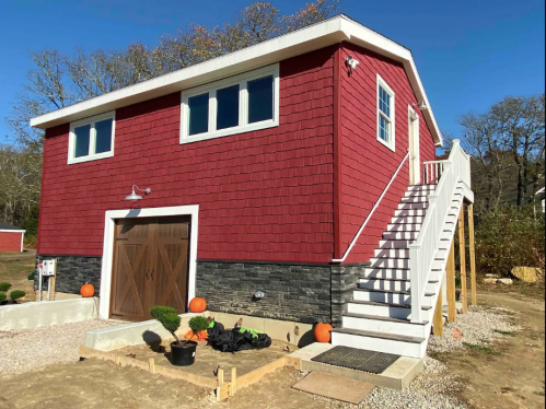 A red two-story house with white trim, stairs, and decorative pumpkins in front, set in a natural landscape.