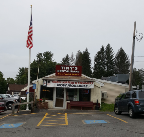 A small restaurant with a sign reading "Tiny's Restaurant" and an American flag, surrounded by trees and parking spaces.
