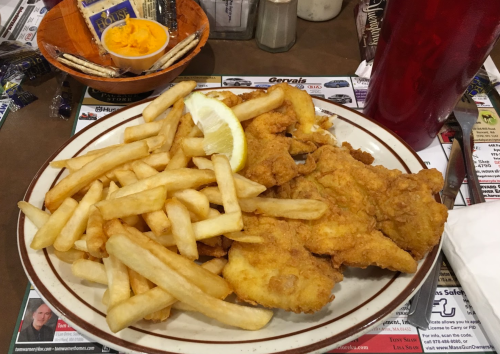 A plate of fried fish fillets with a lemon wedge, served alongside golden French fries and a small bowl of orange sauce.