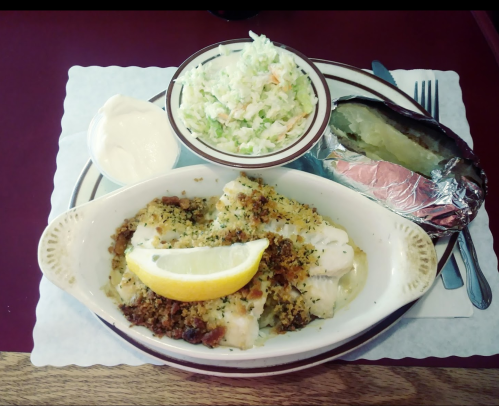 A plate of baked fish topped with breadcrumbs, a lemon wedge, coleslaw, and a baked potato with sour cream on the side.