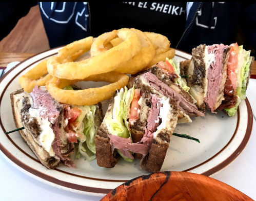 A plate with a stacked sandwich filled with meats, lettuce, and tomatoes, accompanied by crispy onion rings.