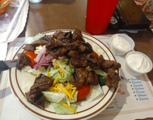 A plate of salad topped with grilled steak, tomatoes, onions, and cheese, accompanied by two cups of dressing.