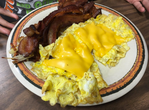 A plate of cheesy scrambled eggs with crispy bacon and hash browns, served on a rustic table.