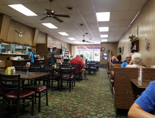 A cozy diner interior with tables, patrons dining, and a server attending to customers. Green carpet and wooden decor.