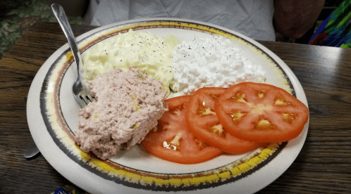 A plate with sliced tomatoes, cottage cheese, salad, and a portion of pink meat spread, served with a fork.
