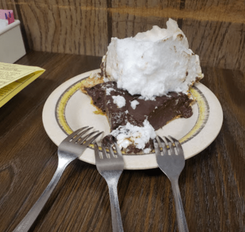 A slice of chocolate pie topped with fluffy meringue, served on a plate with three forks beside it.