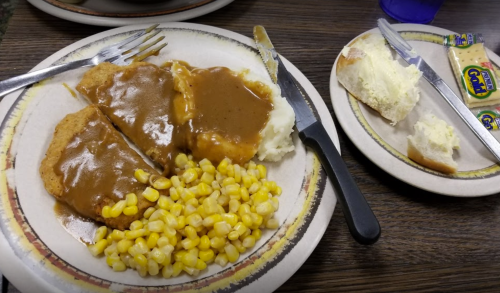 A plate with chicken smothered in gravy, mashed potatoes, and corn, alongside a roll and butter on a separate plate.