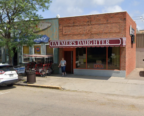A brick storefront with a sign reading "Farmer's Daughter" and an open sign, with a person standing outside.