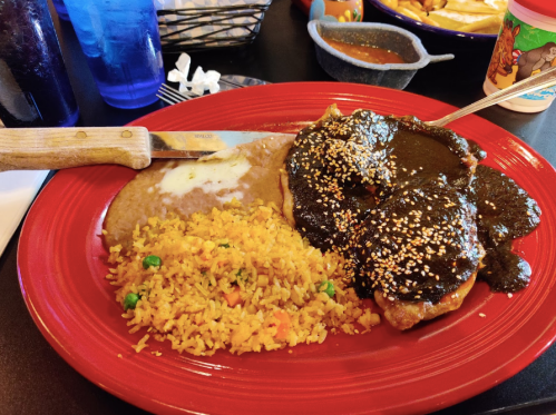 A vibrant red plate with chicken in mole sauce, rice with peas, refried beans, and a knife on the side.