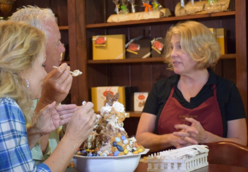 A woman in an apron talks to two people enjoying a large dessert in a cozy shop filled with baked goods.